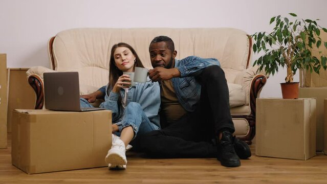 Happy african american man and caucasian woman moving to new home. Newly married couple sitting next to couch drinks tea and talks via video call.