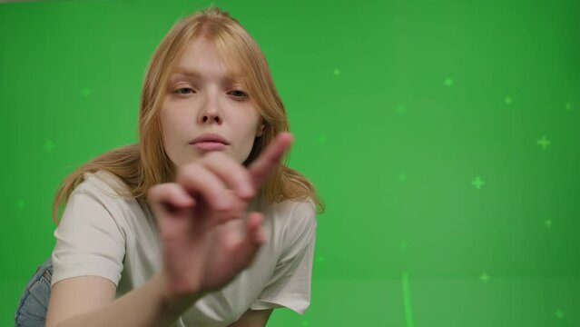 Close-up of Attractive red-haired white girl swipes finger at display of phone. Young woman in jeans and knitted jumper in a positive mood over green screen background, Chroma Key. 4k video footage