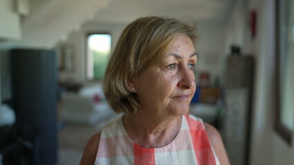 Portrait of a contemplative senior woman standing indoors at home living room looking out window....