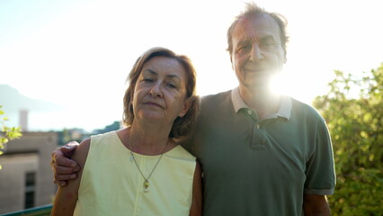 Portrait of a senior couple standing outdoors during sunset posing for camera. An older husband with arm around wife
