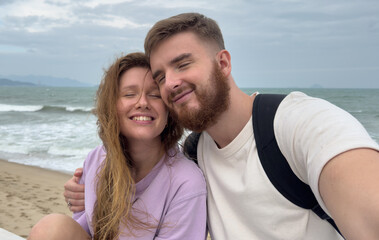 Fototapeta premium Happy couple in love is enjoy summer vacation on sea, beach take selfie photo, picture of themself on phone, smartphone, hug each other 