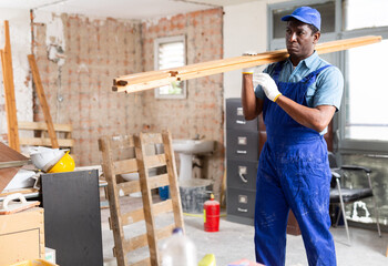 Skilled african american carpenter in blue workwear carrying wooden planks at construction site indoors..