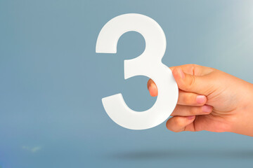Number three in hand. Hand holding white number 3 on blue background with copy space. Concept with...