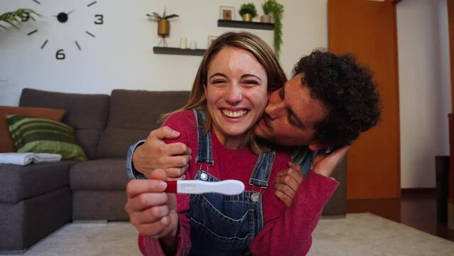 Happy caucasian young couple smiling excited showing at camera their positive pregnancy test at home. Husband and wife celebrating that they are expecting a baby holding a predictor in their hands