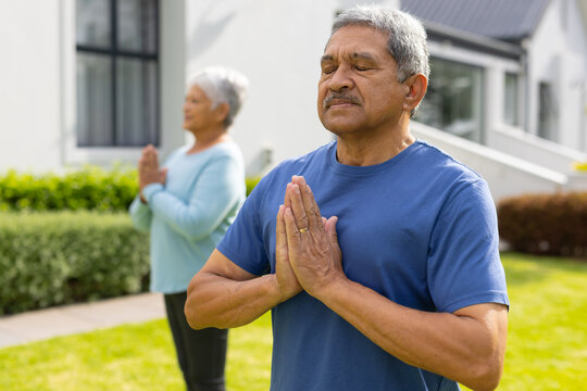 Biracial senior man and woman meditating in prayer pose against house at yard