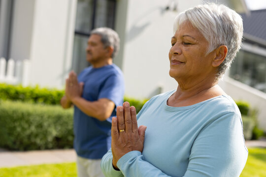 Biracial senior woman and man with eyes closed meditating in prayer pose against house at yard
