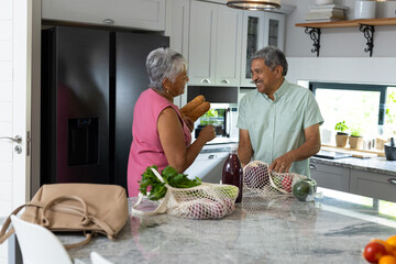 Biracial senior couple talking while unpacking groceries on kitchen counter at home, copy space
