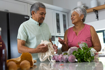 Biracial senior man assisting wife in unpacking groceries from mesh bags on kitchen counter at home