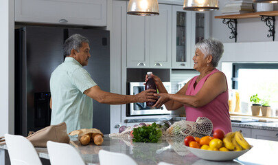 Biracial senior woman giving juice bottle to husband while unpacking groceries on kitchen counter