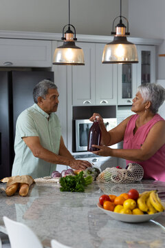 Biracial senior wife giving juice bottle to husband while unpacking groceries on counter in kitchen