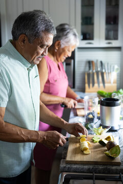 Biracial senior couple cutting fruits into slices on counter while preparing smoothie in kitchen