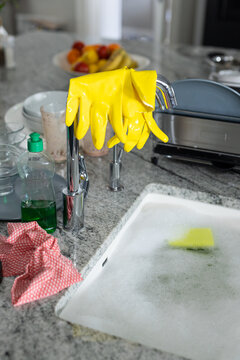 Soap dispenser by yellow washing up gloves on faucet and kitchen sink filled with soapy water