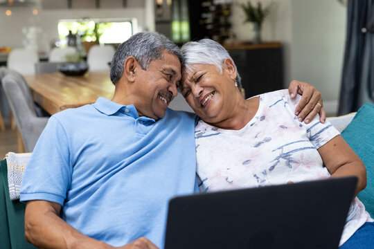 Cheerful biracial senior couple with head to head using laptop while relaxing on sofa - Powered by Adobe