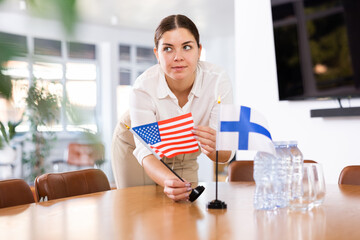 Positive young woman putting little flag of the USA on the table with flag of Finland in conference...