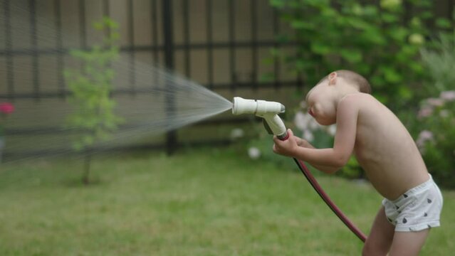 little helping child naked barefoot watering plants holding water hose spray in green garden outdoors in hot summer day. warm weather kid boy working outside developing assistance parenting lifestyle