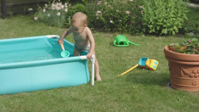 naked caucasian toddler baby boy getting into pool of water outdoors summer day. little child having fun in garden warm sunny day outside. happy kid playing in swimming pool, sitting in water on green