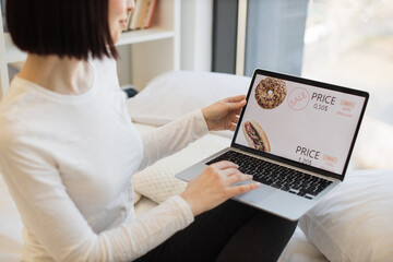 Fototapeta premium Dark haired caucasian woman scrolling website for choosing tasty sweet donuts during discount of 60 percent Casually dressed lady using portable laptop for online shopping.