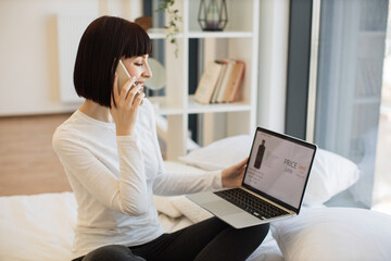 Happy caucasian woman resting on bed with wireless laptop and using smartphone for ordering cosmetic during sale season. Discount of product with 65 percent in online shop.
