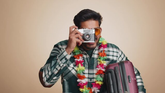 Happy indian man tourist photographer in sunglasses taking pictures photos on retro camera. Travel, summer holiday vacation, trip to seaside. Handsome bearded hindu guy traveler on beige background