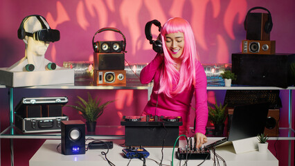 Asian musician playing electronic music at mixer console while having fun dancing in club during night time. Artist with pink hair performing remix sound using professional audio equipment