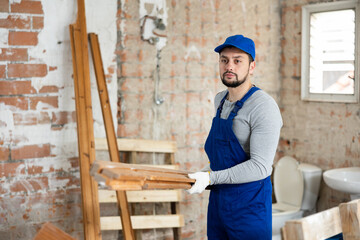 Worker in blue overalls carries planks to renovate a room