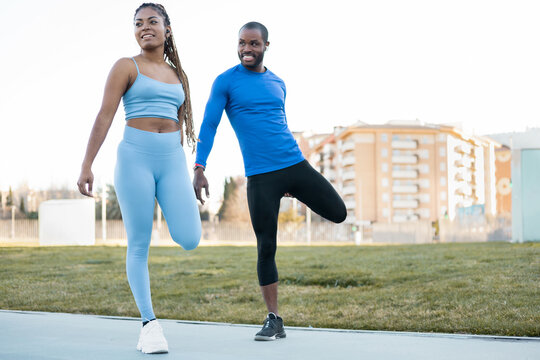 Full length photo of a dark-skinned couple doing sports outdoors together, man and woman are stretching their legs after exercise, the black girl with braids is in the foreground showing movement.