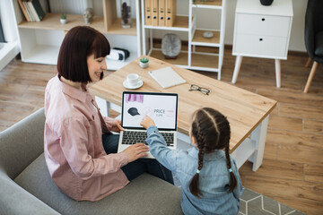 Fototapeta na wymiar Rear view of dark haired woman sitting on couch with laptop on knees while her pretty girl pointing on screen with forefinger. Mother and daughter choosing new clothes to buy during discounts.