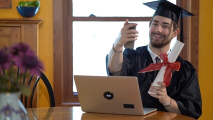 Young man in graduation costume in front of laptop in a home, taking a selfie.