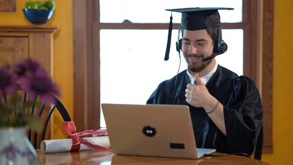 Young man in graduation costume wearing headset with microphone talking to someone on laptop computer.