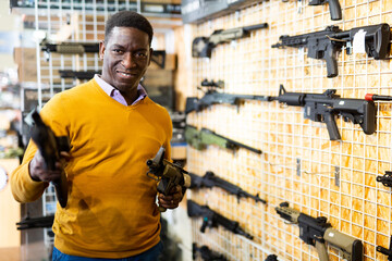 African-american man standing in arms shop and holding two guns in hands.