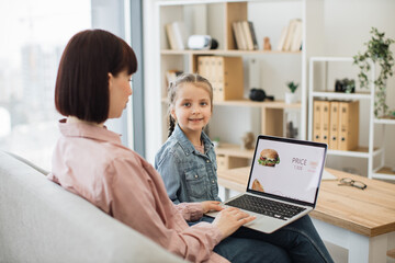Fototapeta na wymiar Side view of charming brunette sitting on couch with daughter and using wireless laptop. Cute little girl smiling and looking at camera while focused mother scrolling online shop with fast food.
