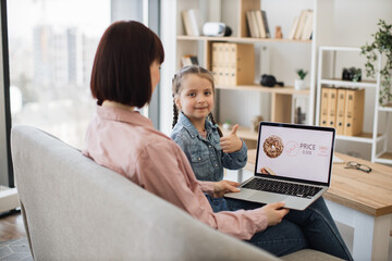 Fototapeta na wymiar Side view of charming brunette sitting on couch with daughter and using wireless laptop. Girl smiling and looking at camera, showing thumb up while focused mother scrolling online shop with fast food.