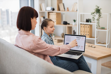 Caring mother with little daughter browsing ecommerce site for searching discount of new trendy apparel. Young woman and pretty girl sitting together on comfy couch and using modern wireless laptop.