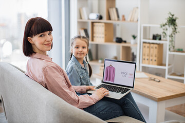 Young mother with daughter resting together on comfy sofa with laptop on knees, smiling and looking at camera. Internet shop offering clothes with discount on computer screen. Online shopping concept.