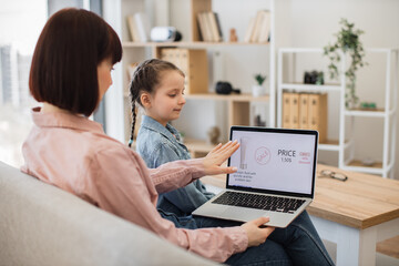 Fototapeta premium Brunette caucasian ladies resting in lovely family room and browsing e-commerce cosmetics webstore using laptop application. Pretty woman and kid searching for online discounts on beauty products.