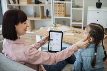 Fototapeta na wymiar Blurred foreground of attractive mother in apartment touching daughter's cheek while holding electonic device in hand. Focus on digital tablet with virtual cosmetics store website offering discounts.