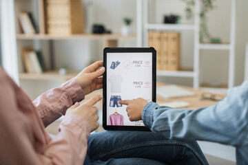 Close up of digital tablet with commercial newcommers of clothes available at discount on screen. Mother and child hanging out in apartment and enjoying internet shopping via electronic gadget.