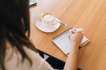 Young businesswoman focused on taking notes in a notebook while sitting at the table with a cup of coffee. Cheerful student sitting at table and taking notes in a notebook
