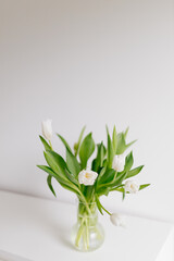 An engagement ring in white gold with a diamond lies in a bouquet of white tulips on white background. Gift for Women's Day, Valentine's Day. Beautiful spring background with green leaves. Mock up