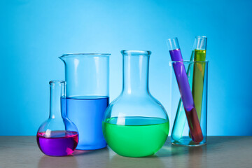 Different laboratory glassware with colorful liquids on wooden table against light blue background