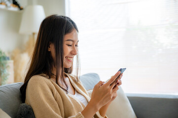 Happy young asian woman relax on comfortable couch at home texting messaging on smartphone, smiling...