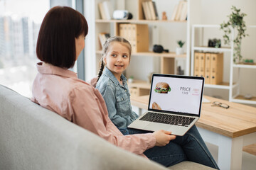 Fototapeta na wymiar Attractive dark haired woman sitting on couch with her lovely daughter and choosing fast food with discount on website. Caucasian family of two using modern laptop for ordering takeaway lunch.