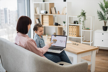 Obraz na płótnie Canvas Attractive dark haired woman sitting on couch with her lovely daughter and choosing fast food with discount on website. Caucasian family of two using modern laptop for ordering takeaway lunch.