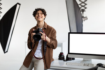 Happy male photographer with professional camera sitting on his desk with computer monitor with blank screen, mockup
