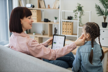 Blurred foreground of attractive mother in apartment touching daughter's cheek while holding electonic device in hand. Focus on digital tablet with virtual cosmetics store website offering discounts.