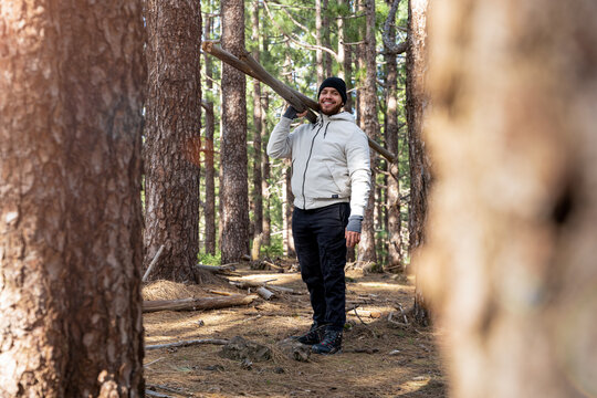 Hombre sonriente cargando troncos de madera en el bosque viendo a c&aacute;mara cuerpo completo