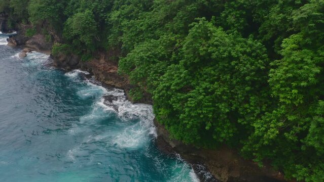Beautiful aerial view of tidal waves splashing on brown rocks with green trees. Amazing copter footage of turquoise sea water washing rocky island. Idyllic tropical scene. Stormy ocean at wild lagoon.