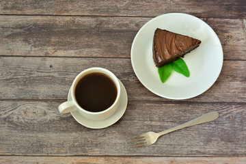 A plate with a piece of mint-decorated chocolate layer cake, a dessert fork and a cup of hot coffee on a saucer on a wooden table.