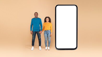 Happy african american spouses standing near huge cellphone with blank screen, advertising application or website, mockup