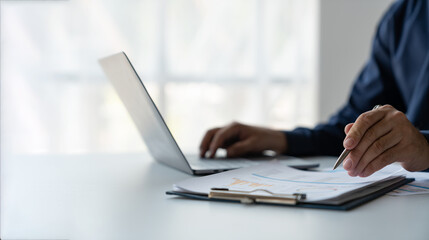 young business man Recording data on laptop, comparing details from graphs. Business income charts in the areas of real estate, tax, management, technology marketing in a private office.